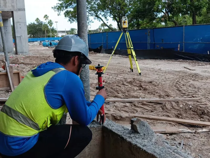 SurveyFy surveyor with a prism pole and total station laying out a foundation on an active construction site